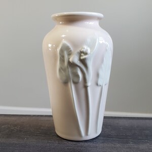 May include: A pale pink ceramic vase with a flared top and a cylindrical body. The vase features three white, raised floral designs. The vase is approximately 10 inches tall.