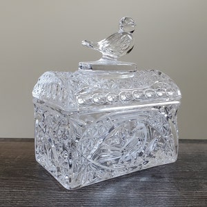 May include: A clear, rectangular crystal trinket box with a bird-shaped lid handle. The box features intricate cut glass patterns and a detailed bird design on the front. The lid is arched and the box is sitting on a dark surface.