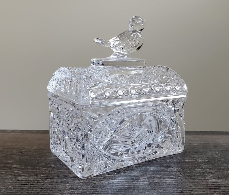 May include: A clear, rectangular crystal trinket box with a bird-shaped lid handle. The box features intricate cut glass patterns and a detailed bird design on the front. The lid is arched and the box is sitting on a dark surface.