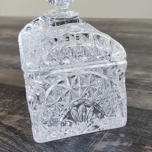 May include: A clear, rectangular crystal trinket box with a lid. The box features a detailed, geometric pattern and a decorative finial on top. The box is sitting on a dark wooden surface.