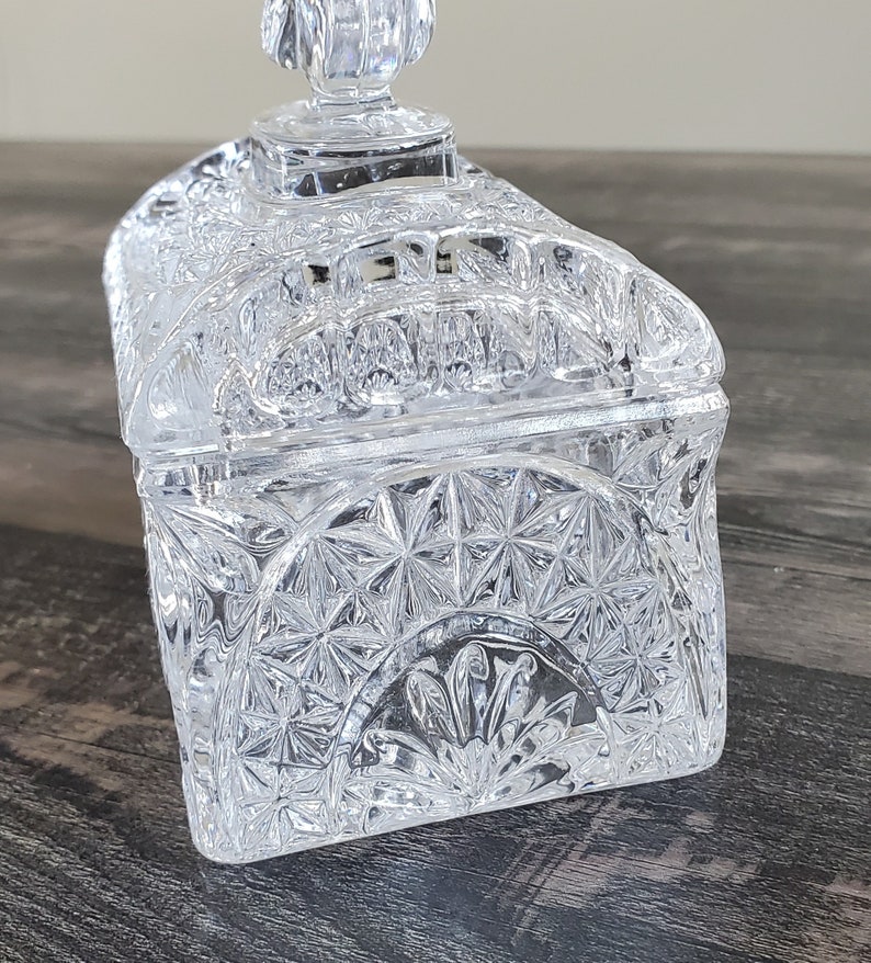 May include: A clear, rectangular crystal trinket box with a lid. The box features a detailed, geometric pattern and a decorative finial on top. The box is sitting on a dark wooden surface.