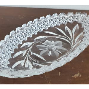 May include: Clear glass oval dish with a floral and leaf design etched into the center. The rim has a repeating geometric pattern. The dish is sitting on a brown wooden surface.