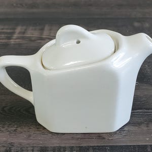 May include: A small, white ceramic teapot with a heart-shaped lid. The teapot has a handle and a spout. The lid has a small hole. The teapot is on a dark wooden surface.