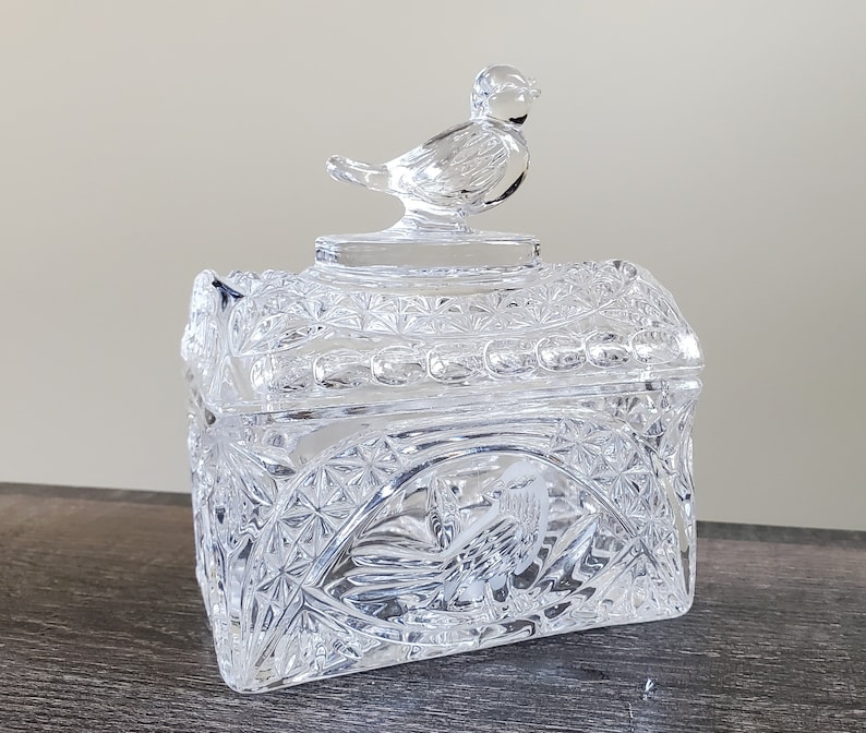 May include: A clear, square-shaped crystal trinket box with a bird-shaped lid. The box features intricate geometric and floral patterns. The lid has a small, detailed bird figurine. The box is displayed on a dark wooden surface.
