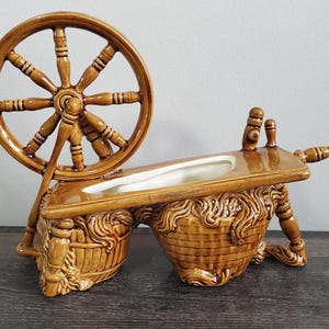May include: A brown ceramic spinning wheel planter. The planter features a large wheel, a rectangular trough, and a decorative base with a basket weave design. The planter is a decorative item.
