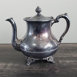 May include: A silver-colored teapot with a curved spout and ornate handle. The teapot has a lid with a decorative finial and sits on a detailed base. The body of the teapot features engraved floral patterns.