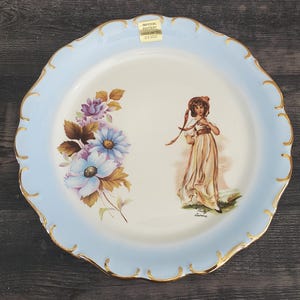 May include: A decorative plate with a light blue scalloped edge and gold trim. The plate features a floral design with blue and purple flowers and a woman in a long dress. The plate has a label that says "Imperial Pottery".