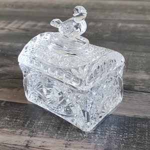 May include: A clear, cut-glass trinket box with a bird-shaped lid handle. The box is rectangular with intricate geometric patterns and a decorative bird on top. The box is made of clear glass and sits on a wooden surface.