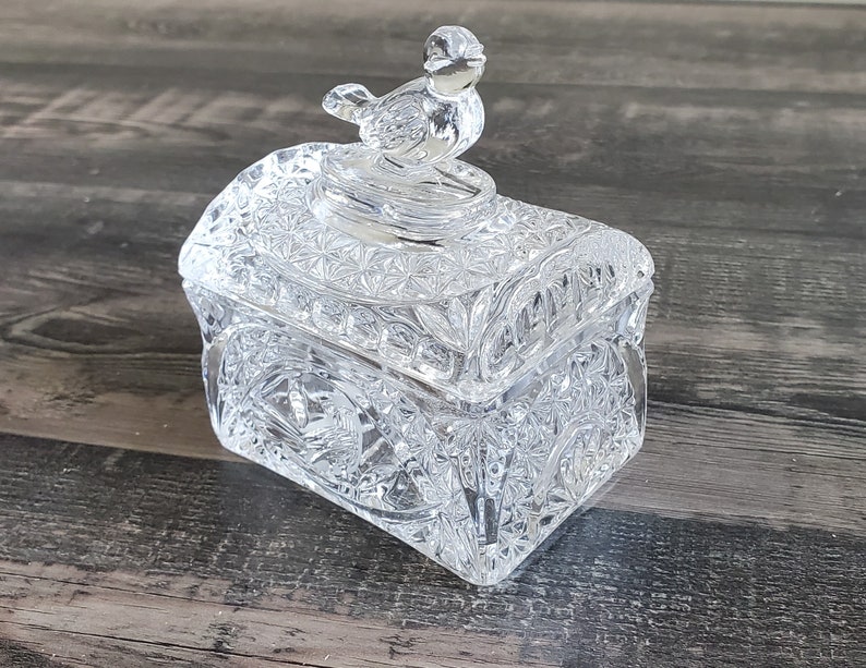 May include: A clear, cut-glass trinket box with a bird-shaped lid handle. The box is rectangular with intricate geometric patterns and a decorative bird on top. The box is made of clear glass and sits on a wooden surface.