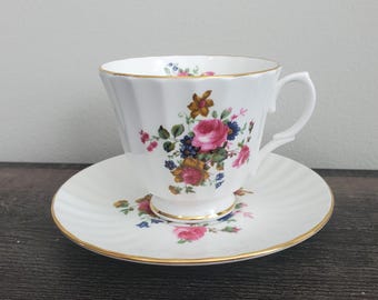 May include: A white teacup and saucer set with a floral pattern. The cup has a fluted rim and a delicate handle. The saucer has a scalloped edge and a gold trim.