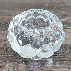 May include: A clear glass votive candle holder with a textured, bubble-like design. The candle holder is round with a central opening and sits on a wooden surface. The glass is transparent, allowing light to pass through.