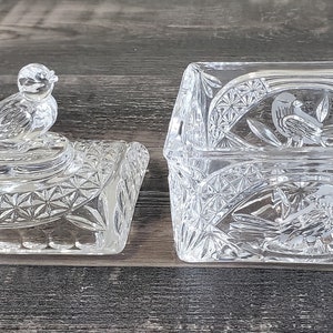 May include: A clear, cut-glass trinket box with a matching lid. The lid features a bird figurine. The box and lid have a geometric pattern and bird motifs. The box is rectangular and sits open on a wooden surface.