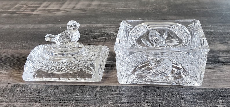 May include: A clear, cut-glass trinket box with a matching lid. The lid features a bird figurine. The box and lid have a geometric pattern and bird motifs. The box is rectangular and sits open on a wooden surface.