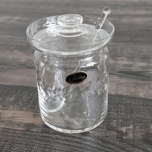 May include: A clear glass jar with a lid and a small spoon. The jar has a textured pattern and a small label that reads "Cambridge". The spoon rests on the lid. The jar is sitting on a wooden surface.