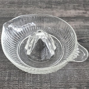 May include: Clear glass citrus juicer with a ribbed bowl and a central reamer. The juicer has a handle for easy pouring. The design is simple and functional, perfect for squeezing fresh juice.