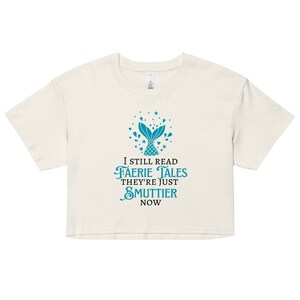 May include: A white cropped t-shirt with a teal mermaid tail graphic and the text "I still read faerie tales they're just smuttier now".