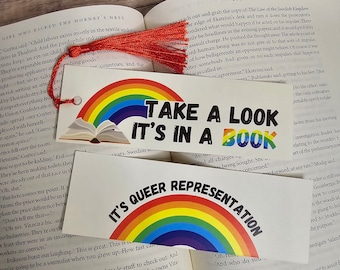 Puede incluir: Dos marcadores con diseños de arcoíris y texto. Uno dice "TAKE A LOOK IT'S IN A BOOK" y el otro "IT'S QUEER REPRESENTATION". Una borla roja cuelga del marcador superior, apoyada en un libro abierto.