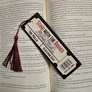 Op de afbeelding: Een zwart-witte bladwijzer met de tekst "STAND WITH THE BANNED Book Club ELECTION DAY" en een barcode. Het heeft een bordeauxrode kwast. De bladwijzer ligt op een open boek.