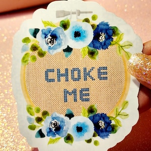 May include: A white sticker with a blue floral border and a tan cross-stitch design in the center. The cross-stitch design says "CHOKE ME".