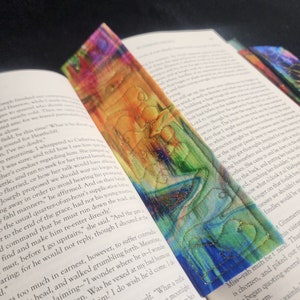 May include: A colorful bookmark with a quilted design and gold thread embroidery. The bookmark features a vibrant abstract pattern with shades of blue, green, yellow, orange, and red.