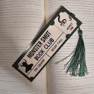 May include: A green bookmark with a black and white illustration of a monster. The bookmark has the text "Monster Smut Book Club" and "The Swamp" printed on it. The bookmark also has a green tassel.