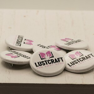 以下が含まれることがあります： 「Lustcraft The Smut Shop」と書いてある黒とピンクのロゴがついた白い丸いボタン6個のクローズアップ。