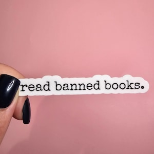 May include: A white sticker with the text "read banned books." The sticker is being held by a hand with black nail polish.
