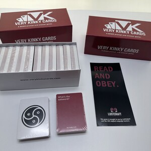 May include: A set of Very Kinky Cards, a blush prompt and answer game, in a white box with the website address printed on the front. The box is open and the cards are visible inside. There are two red boxes with the same title, "Very Kinky Cards", and a black card with the text "Read and Obey." There are two cards on the table, one with a symbol and the other with the text "What's the safeword?"
