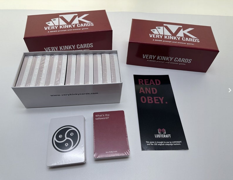 May include: A set of Very Kinky Cards, a blush prompt and answer game, in a white box with the website address printed on the front. The box is open and the cards are visible inside. There are two red boxes with the same title, "Very Kinky Cards", and a black card with the text "Read and Obey." There are two cards on the table, one with a symbol and the other with the text "What's the safeword?"