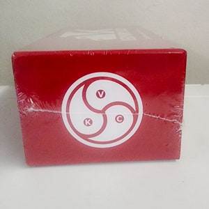 May include: Red box with a white circle containing a black triangle with the letters V, K, and C inside. The box is wrapped in clear plastic.