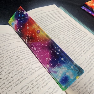 May include: A colorful bookmark with a galaxy print. The bookmark is made of fabric and has a quilted design. The bookmark is folded in half and is lying on top of an open book.