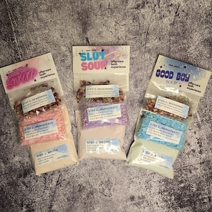 May include: Three packages of bath salts with different labels. The labels are pink, purple, and blue. The pink label says "Good Girl Soup" and has a pink and white bath salt mixture. The purple label says "Slut Soup" and has a purple and white bath salt mixture. The blue label says "Good Boy Soup" and has a blue and white bath salt mixture.