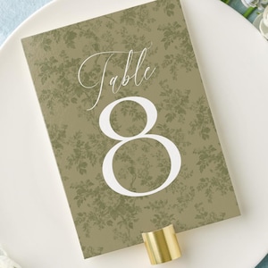 May include: A table setting with a white plate, floral arrangement, and vintage silverware tied with a ribbon. A table number card with the number 8 is displayed on the plate. The background is a light blue painted surface.