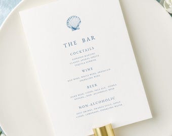 Pode incluir: Um prato branco com um cartão de menu "The Bar" com um logotipo de concha, listando coquetéis, vinho, cerveja e opções não alcoólicas. Um porta-guardanapo dourado segura o menu. Garfo e colher vintage amarrados com uma fita branca. Flores azuis e brancas.