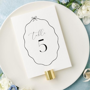 May include: A white plate with a white card on top. The card has a black oval frame with the text "Table 5" inside. The plate is surrounded by blue and white flowers.