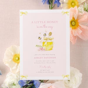 Pode incluir: Um convite para um chá de bebê rosa e amarelo com uma ilustração em aquarela de um pote de mel e abelhas. O texto diz "Um pouco de mel está a caminho" e inclui o nome Ashley Davidson.