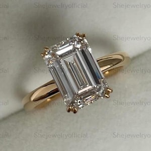 May include: A gold engagement ring with a large, rectangular, clear gemstone. The ring is set in a gold band and is displayed in a green jewelry box. The text "Shejewelryofficial" is visible.