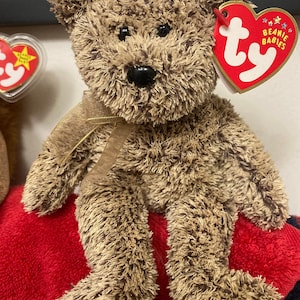 May include: A brown and tan fuzzy Beanie Baby teddy bear with a gold ribbon around its neck. The bear is sitting on a red blanket. The bear has a black nose and black eyes. The bear has a Ty Beanie Babies tag attached to its ear.