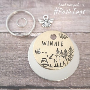 May include: A round pet tag made of gold-toned metal with the name "WINNIE" engraved on it. The tag features a black and white illustration of a bear, a bee, and a honeycomb. The tag is attached to a silver key ring with a silver bee charm.