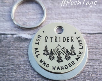 May include: A round, silver, double-sided key chain with a brass tag. The brass tag has a black stamped design of a mountain range with trees and the text "STRIDER NOT ALL WHO WANDER ARE LOST".