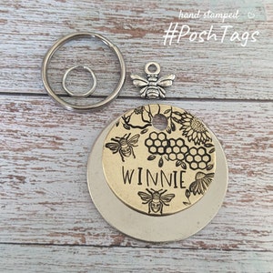 May include: A gold-toned metal pet tag featuring a beehive design and the name "WINNIE" engraved on it. The tag is attached to a silver key ring with a small bee charm.