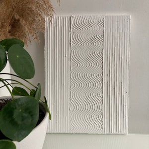 Könnte beinhalten: Weiße strukturierte Leinwandkunst mit drei Abschnitten. Die linken und rechten Abschnitte haben vertikale Linien, und der mittlere Abschnitt hat ein wellenförmiges Muster.