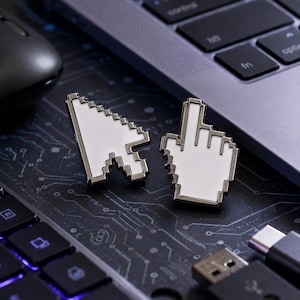 May include: Two enamel pins shaped like computer mouse cursors. One pin is a white arrow pointer with a dark metal outline, and the other is a white pointing hand icon with a matching dark metal border, both featuring a pixelated design.