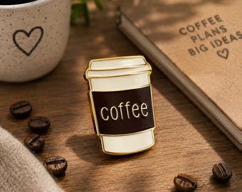 May include: A gold-toned enamel pin shaped like a disposable coffee cup with a white lid and base, featuring a dark brown center band with the word coffee written in gold lettering. The pin has a smooth, glossy finish and a metallic gold border.