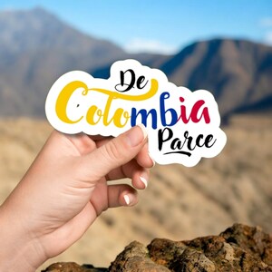 May include: A white sticker with the words "De Colombia Parce" in black, yellow, blue, and red. The sticker is held in a person's hand against a blurred background of mountains and a blue sky.