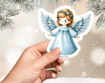 May include: A sticker featuring a cartoon angel with light blue wings and a matching dress. The angel has blonde hair and a green floral crown. The sticker is held in a hand, with the text "designs & Art" visible.