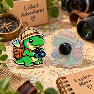 May include: Two acrylic pins featuring a cartoon green dinosaur explorer wearing a tan hat, carrying a brown backpack with a map, and holding black binoculars. One pin shows the front design, while the other shows the black metal pin back.