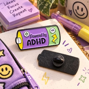 May include: A battery-shaped enamel pin featuring a purple center with the text Powered by ADHD in black, a yellow end, and a light green end with a yellow lightning bolt. The pin includes a black metal backing with a rubber clutch fastener.
