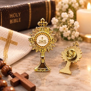 May include: Two gold-toned metal monstrance pins featuring a central white circular emblem with the letters JHS and a small cross. One pin is displayed from the front, showing the detailed sunburst design, while the other shows the back metal clasp.