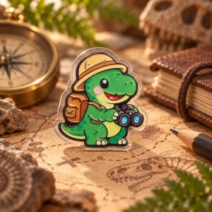 May include: A clear acrylic pin featuring a cartoon green dinosaur wearing a tan explorer hat and a brown backpack while holding black binoculars. The dinosaur has a light green belly, pink cheeks, and a wide smile, rendered in a flat illustration style.